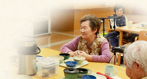 食堂で食事をする高齢者入居者の風景。職員も同席しており、温かい食事風景が捉えられている。